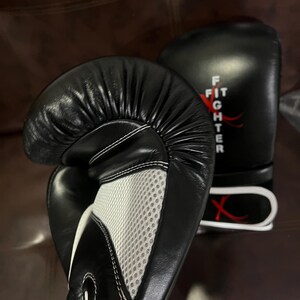 May include: A pair of black boxing gloves featuring a white mesh palm panel and white stitching. The word FIGHTER is printed vertically in white on the wrist area, accompanied by a red graphic design. The gloves have a smooth, padded surface.