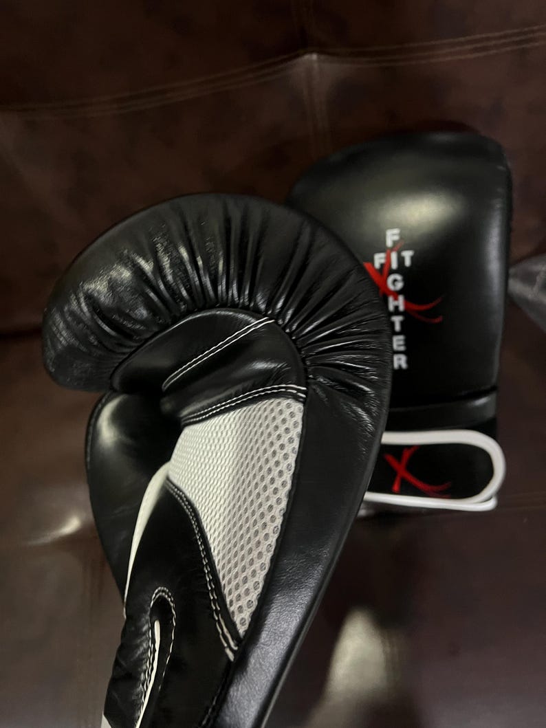 May include: A pair of black boxing gloves featuring a white mesh palm panel and white stitching. The word FIGHTER is printed vertically in white on the wrist area, accompanied by a red graphic design. The gloves have a smooth, padded surface.