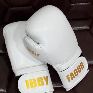 May include: A pair of white boxing gloves featuring gold lettering. One glove displays the word WOLF on the side and IBBY on the wrist strap, while the other glove features the name FAOUR in gold on the white wrist strap closure.