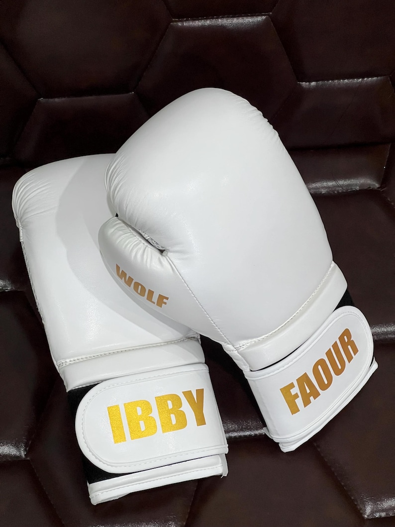 May include: A pair of white boxing gloves featuring gold lettering. One glove displays the word WOLF on the side and IBBY on the wrist strap, while the other glove features the name FAOUR in gold on the white wrist strap closure.
