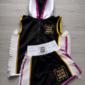 May include: A black sleeveless hooded vest and matching shorts set featuring white fringe trim, gold piping, and metallic magenta accents. The garments display the text YOUR LOGO HERE in gold on the chest, waistband, and leg, with a white hood lining.