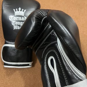 May include: A pair of black boxing gloves with white trim and stitching. One glove features a white crown logo above the text Tornado Tong Ho. The gloves have a padded wrist area and a secure closure system, showcasing a smooth, durable exterior finish.
