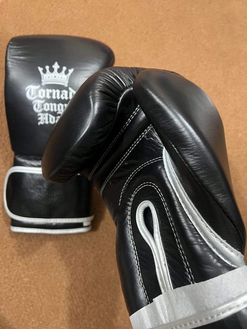 May include: A pair of black boxing gloves with white trim and stitching. One glove features a white crown logo above the text Tornado Tong Ho. The gloves have a padded wrist area and a secure closure system, showcasing a smooth, durable exterior finish.
