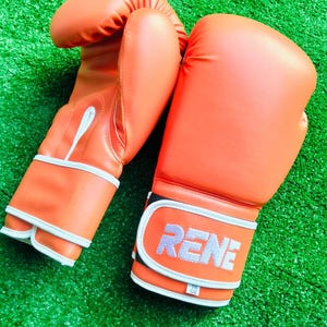 May include: A pair of bright orange boxing gloves with white trim and piping. The wrist strap features the text RENE in white lettering, with a small white tag below it displaying 16 oz. The gloves have a smooth, solid-colored exterior finish.