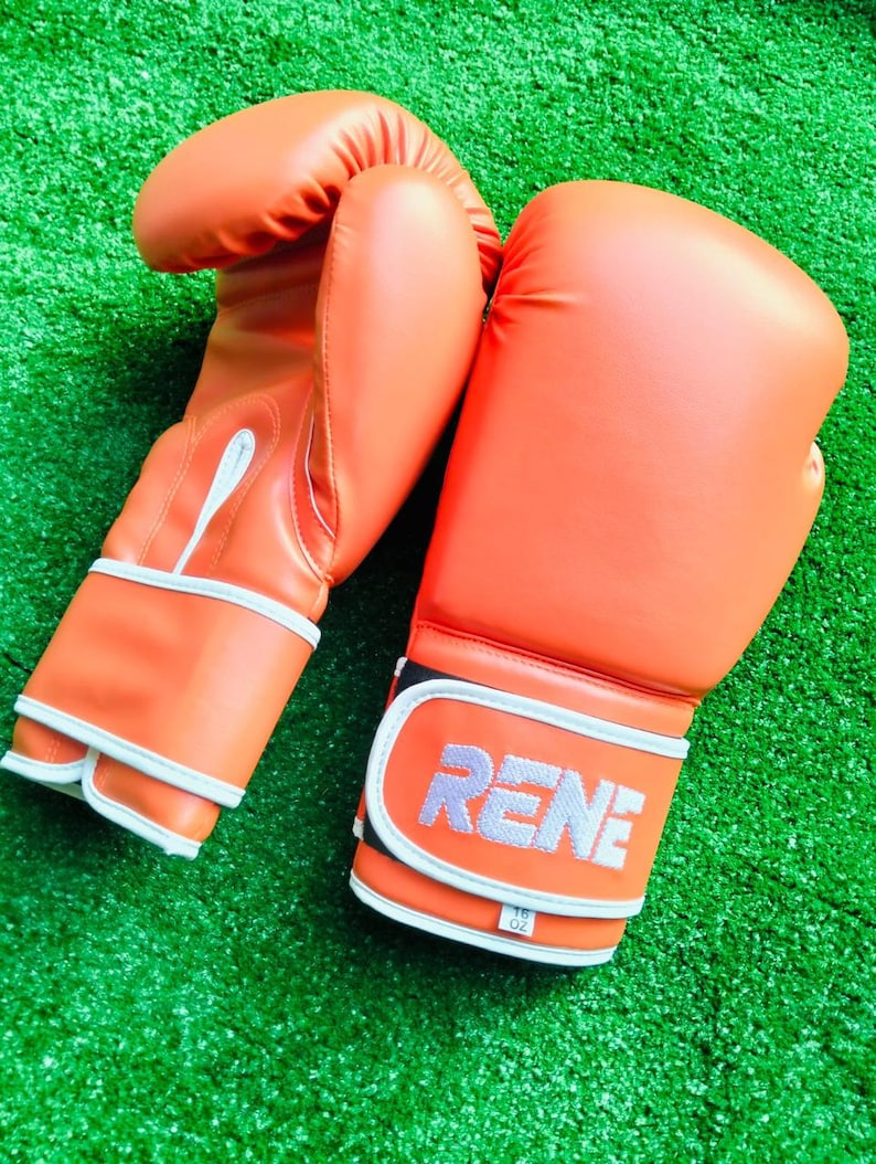 May include: A pair of bright orange boxing gloves with white trim and piping. The wrist strap features the text RENE in white lettering, with a small white tag below it displaying 16 oz. The gloves have a smooth, solid-colored exterior finish.