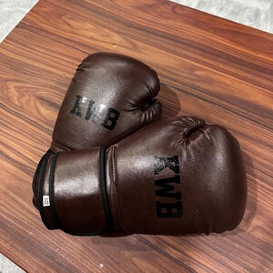May include: A pair of dark brown boxing gloves featuring the black text KWB printed on the front of each glove. The wrist area of one glove displays a small white tag with the text 8-OZ, indicating the weight of the equipment in ounces.