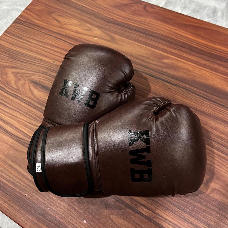 May include: A pair of dark brown boxing gloves featuring the black text KWB printed on the front of each glove. The wrist area of one glove displays a small white tag with the text 8-OZ, indicating the weight of the equipment in ounces.