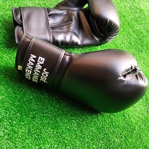 May include: A pair of black boxing gloves with a smooth finish. The wrist area of one glove features the white printed text JOS&Eacute; EMMANUEL MAKENGO. The gloves have a padded thumb section and a secure wrist strap closure system for support during use.