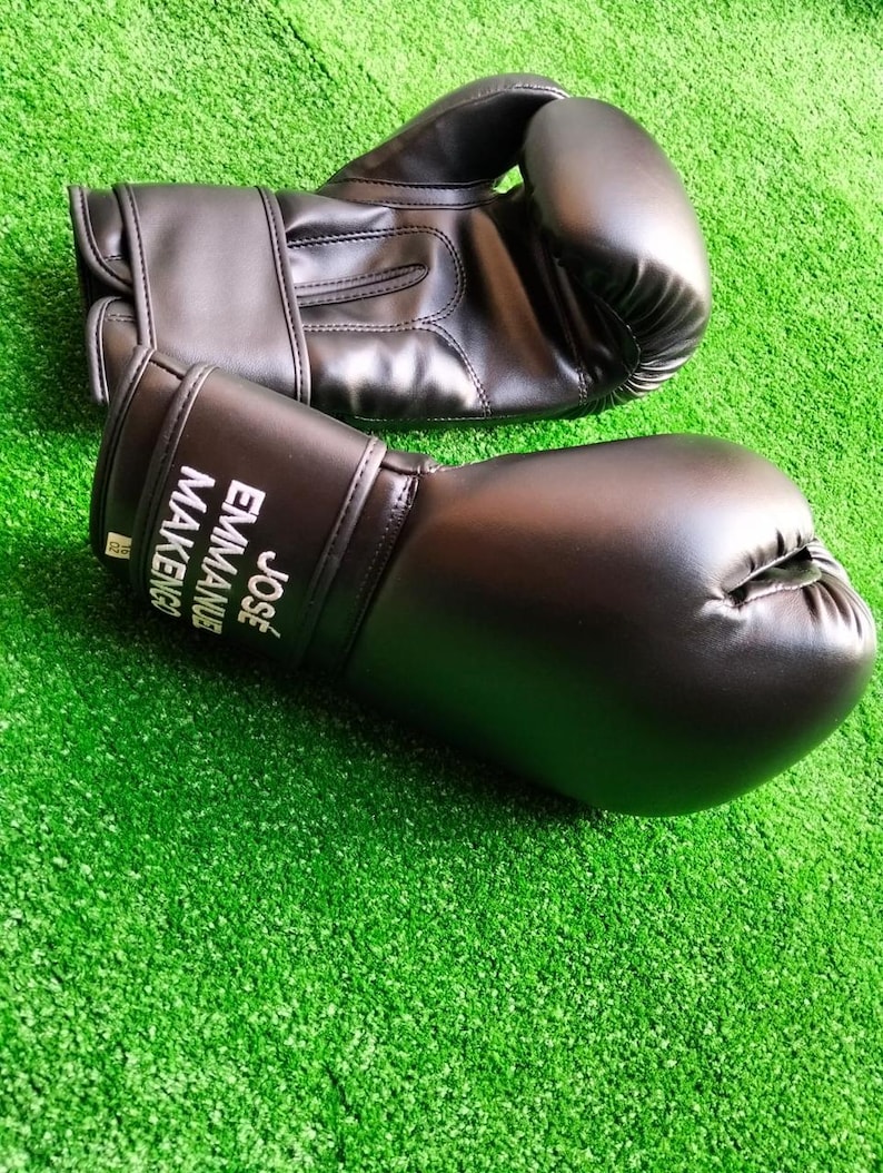 May include: A pair of black boxing gloves with a smooth finish. The wrist area of one glove features the white printed text JOS&Eacute; EMMANUEL MAKENGO. The gloves have a padded thumb section and a secure wrist strap closure system for support during use.