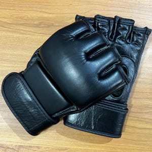 Puede incluir: Un par de guantes de MMA negros de cuero. Los guantes tienen dedos abiertos y nudillos acolchados para protección. La zona de la muñeca también está acolchada para soporte. Estos guantes están diseñados para el entrenamiento y la competición de artes marciales mixtas.