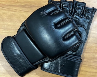 May include: A pair of black fingerless combat gloves featuring a smooth, padded knuckle section and a textured leather-like finish. The gloves include a wide, adjustable wrist strap with a secure closure system and open finger slots for mobility.