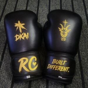 May include: A pair of black boxing gloves with yellow graphics. The left glove features a cross symbol above the text DKAI and the letters RC with a heart. The right glove displays a goat head with a cross and the text BUILT DIFFERENT.