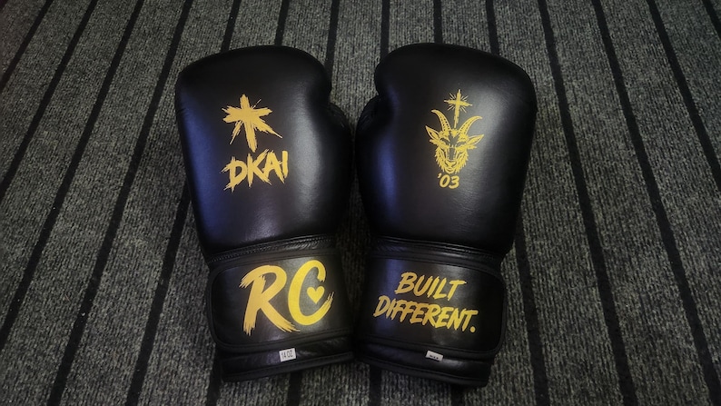 May include: A pair of black boxing gloves with yellow graphics. The left glove features a cross symbol above the text DKAI and the letters RC with a heart. The right glove displays a goat head with a cross and the text BUILT DIFFERENT.