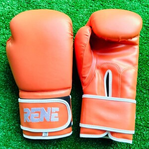 May include: A pair of bright orange boxing gloves with white trim and a white wrist strap. The left glove features the word RENE in white block letters on the strap, with a small white tag below it labeled 16 OZ. The right glove has a ventilation slit.