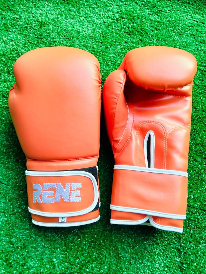 May include: A pair of bright orange boxing gloves with white trim and a white wrist strap. The left glove features the word RENE in white block letters on the strap, with a small white tag below it labeled 16 OZ. The right glove has a ventilation slit.