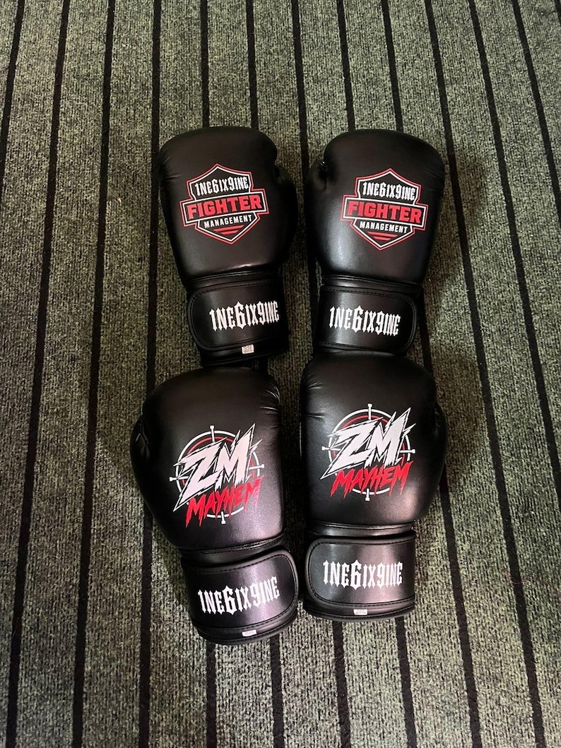 May include: Two pairs of black boxing gloves. The top pair features a red and white shield logo with the text 1NEGIX9INE FIGHTER MANAGEMENT. The bottom pair displays a white and red ZM MAYHEM graphic. All wrist straps are printed with 1NEGIX9INE.