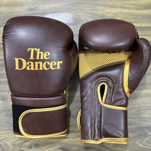 May include: A pair of brown boxing gloves with gold trim and gold mesh detailing on the palm side. The left glove features the text "The Dancer" printed in gold lettering on the front. Each glove has a wide wrist strap with a black hook-and-loop closure.