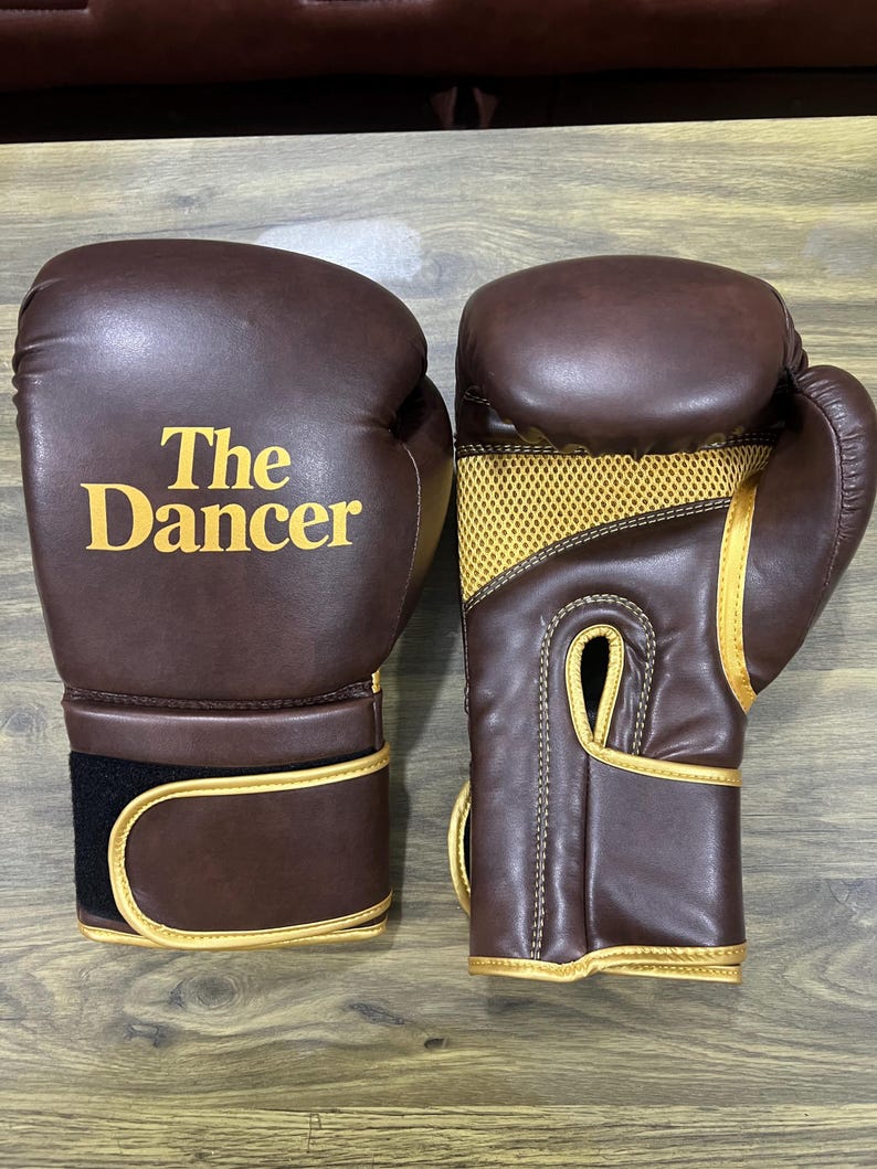 May include: A pair of brown boxing gloves with gold trim and gold mesh detailing on the palm side. The left glove features the text "The Dancer" printed in gold lettering on the front. Each glove has a wide wrist strap with a black hook-and-loop closure.