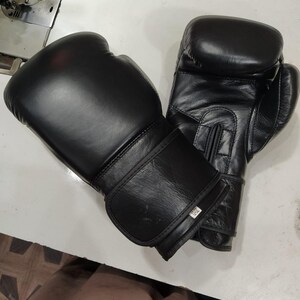 May include: A pair of black boxing gloves with a smooth finish and a hook-and-loop wrist closure. One glove features a small white rectangular tag on the wrist strap with the text 12oz printed in black, indicating the weight in ounces.