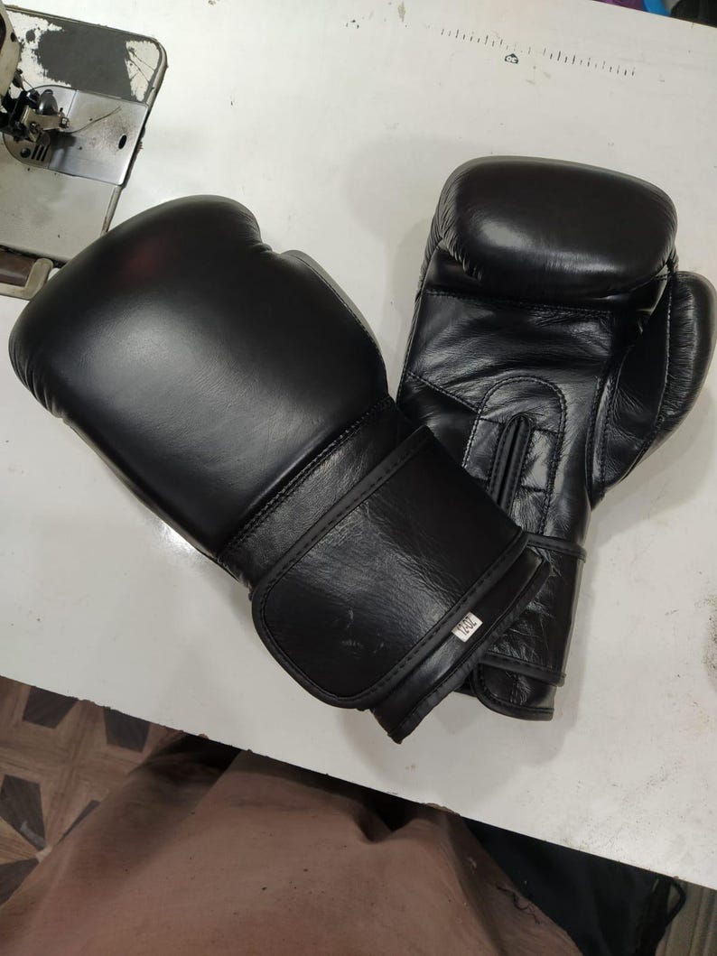 May include: A pair of black boxing gloves with a smooth finish and a hook-and-loop wrist closure. One glove features a small white rectangular tag on the wrist strap with the text 12oz printed in black, indicating the weight in ounces.