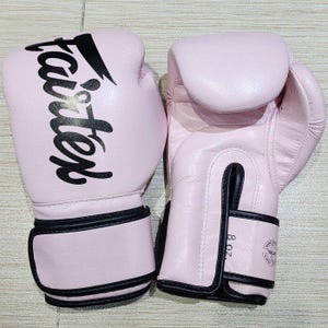 May include: A pair of light pink boxing gloves featuring the black Fairtex logo printed on the front. The gloves have black trim along the wrist straps and a visible 8 oz. weight label printed on the side of the wrist closure.