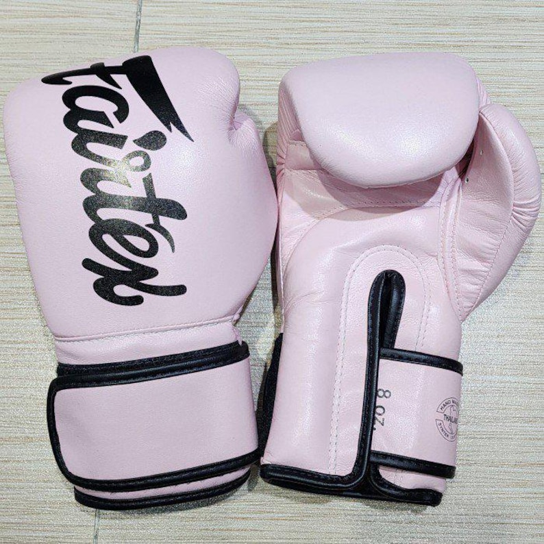 May include: A pair of light pink boxing gloves featuring the black Fairtex logo printed on the front. The gloves have black trim along the wrist straps and a visible 8 oz. weight label printed on the side of the wrist closure.