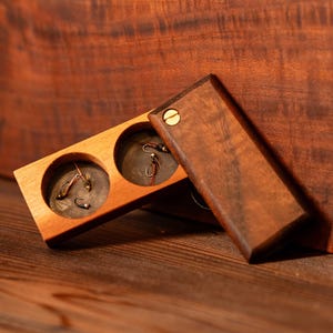 Może przedstawiać: Drewniana skrzynka z klapką, ukazująca dwa przedziały zawierające przynęty wędkarskie. Skrzynka wykonana jest z ciemnego drewna o naturalnym usłojeniu.