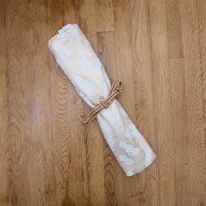 Op de afbeelding: Gerolde witte en goudkleurige stof met patroon, vastgezet met een bruine en bruingele geweven stropdas. De stof heeft een herhalend zeshoekig ontwerp. Het item wordt weergegeven op een houten oppervlak.