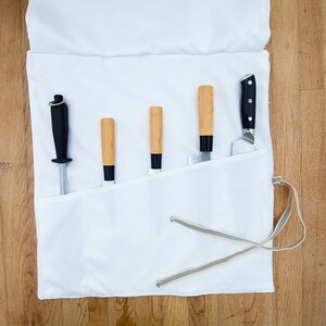 May include: A white canvas knife roll containing five knives and a sharpening steel. The knives have wooden handles and black accents. The roll is open, displaying the tools, and has a drawstring closure.
