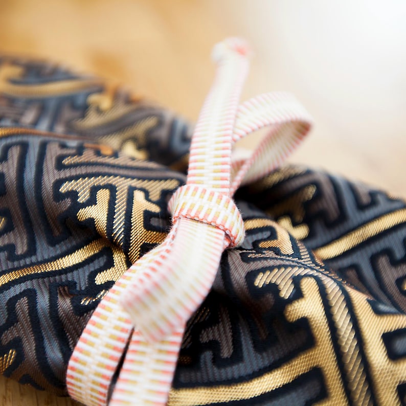 May include: Close-up of a fabric pouch with a geometric pattern in gold, grey, and black. A pink and white striped ribbon is tied in a bow around the pouch. The background is blurred, creating a soft focus.
