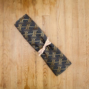 Op de afbeelding: Opgewreven stof met een herhalend goud- en marineblauw patroon, vastgemaakt met een rood-wit gestreept lint. De stof ligt op een houten oppervlak.