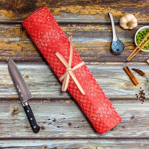 May include: A red patterned knife roll tied with a beige ribbon, next to a chef's knife with a black handle. The scene includes a spoon, chopsticks, and small bowls with ingredients on a wooden surface.