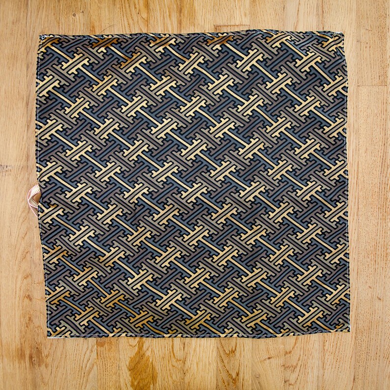 May include: A square textile featuring a repeating geometric pattern in blue, grey, and gold. The fabric is displayed on a wooden surface, highlighting the detailed design. The pattern is a complex arrangement.