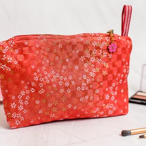 Könnte beinhalten: Rote Kosmetiktasche mit floralem Muster und Karodesign. Die Tasche hat einen Reißverschluss mit einem rosa Blumenanhänger und einem gestreiften Griff. Make-up-Pinsel und eine Palette befinden sich im Hintergrund.
