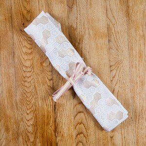 May include: A rolled-up white and beige patterned fabric tied with a pink and white striped ribbon. The fabric features a repeating hexagonal design. The item is resting on a wooden surface.