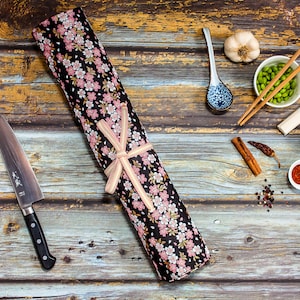 May include: A Japanese chef's knife with a black handle and a floral knife roll. The roll is black with pink and white flowers and tied with a beige ribbon. Other items include a spoon, chopsticks, and small bowls with food.