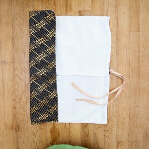 May include: A partially rolled-up white and patterned fabric case with a gold, black, and grey geometric design. The case has a white interior and is tied with a thin, striped cord. The case is on a wooden surface.
