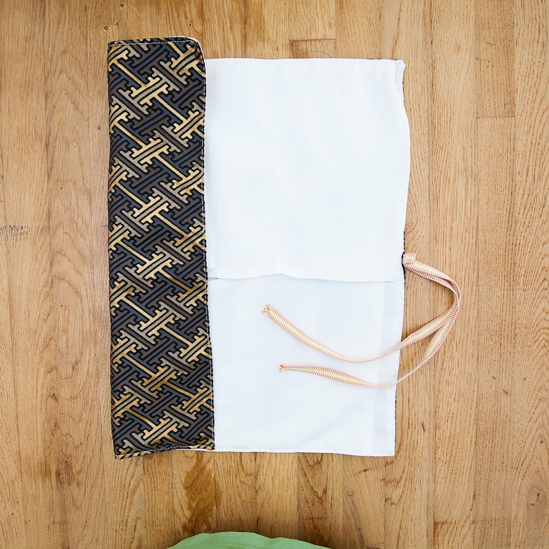 May include: A partially rolled-up white and patterned fabric case with a gold, black, and grey geometric design. The case has a white interior and is tied with a thin, striped cord. The case is on a wooden surface.