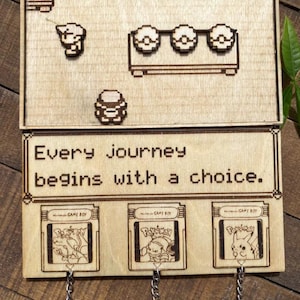 以下が含まれることがあります： ピクセル化されたビデオゲームをテーマにした木製のキーホルダー。キーホルダーは、ゲーム要素のある上部、テキスト「Every journey begins with a choice」、ゲームカートリッジのデザインの3つのキーチェーンが特徴です。キーチェーンはシルバーリングです。