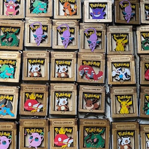 May include: A selection of wooden Pokemon pins, each set within a miniature Game Boy frame. The pins feature various colourful Pokemon characters, with the "Pokemon" and "Nintendo Game Boy" logos visible.