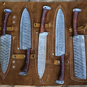 Puede incluir: Un juego de cinco cuchillos de acero de Damasco con mangos de madera, exhibidos en una funda de cuero marrón. Las hojas tienen un patrón texturizado único. La funda tiene correas de cuero y está sobre una superficie de madera. El logotipo "POCKET BLADES" es visible.