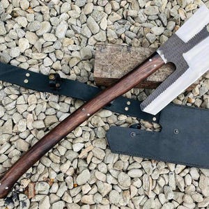 Könnte beinhalten: Eine große Axt aus Metall mit einer breiten, flachen, silberfarbenen Klinge, die eine gezackte Kante und einen kreisförmigen Ausschnitt aufweist. Der Griff besteht aus dunkelbraunem Holz mit sichtbaren Metallnieten. Eine schwarze Lederscheide.