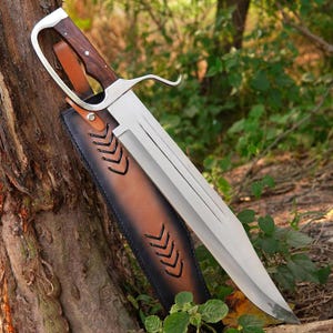 Könnte beinhalten: Ein großes Messer mit feststehender Klinge aus poliertem Stahl, einem Griff aus dunklem Holz und einem Handschutz aus silberfarbenem Metall. Das Messer steckt in einer braun-schwarzen Lederscheide mit Ziernähten und einem beigen Lederriemen.