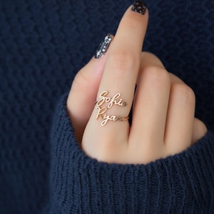 May include: A gold-coloured ring featuring the names "Sofia" and "Ryan" in a cursive font. The ring is worn on a finger, with the names displayed horizontally. The background is a dark blue.