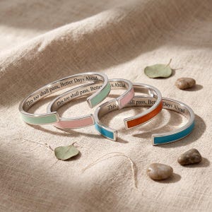May include: Four open cuff bracelets in silver-tone metal, each featuring a colored enamel inlay in mint green, light pink, soft pink, and bright blue. The interior of each bracelet is engraved with the text, This too shall pass, Better Days Ahead.