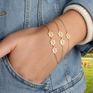 May include: Gold-colored bracelets with paw print charms. The bracelets are layered and worn on a wrist, with a denim jacket in the background. The charms are in the shape of a paw print.
