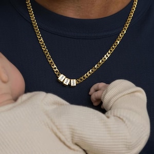Puede incluir: Un collar de cadena dorada con tres pequeños colgantes cúbicos blancos. Cada colgante tiene un nombre grabado en negro. El collar se lleva sobre una camisa azul oscuro. Un bebé está en primer plano.