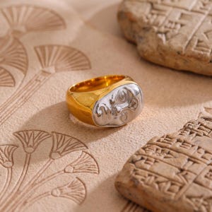May include: A gold-toned metal ring featuring a prominent, raised silver-toned face design on the front. The band is smooth and polished, contrasting with the detailed, sculpted facial features that include eyes, a nose, and lips in a metallic finish.