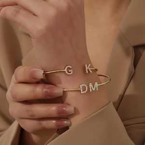 May include: Two gold-colored, open cuff bracelets. One bracelet has the letter "G" and "K" in a sparkling, rhinestone-like font. The other bracelet has the letters "DM" in a similar font. The bracelets are worn on a wrist.