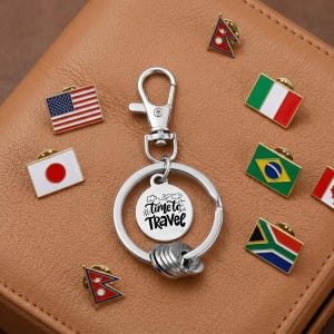 May include: A silver metal keyring featuring a circular charm engraved with the text Time to Travel and a lobster clasp. Arranged around the keyring are multiple small enamel lapel pins displaying various national flags, including those of Japan and the USA.